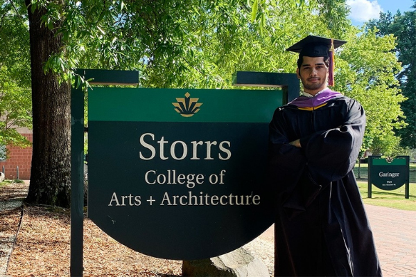 photo of recent graduate in cap and gown in front of the storrs sign