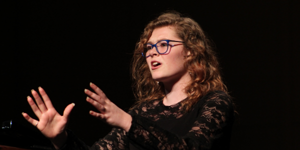 woman in glasses sining with her hand open in front of her against a black stage