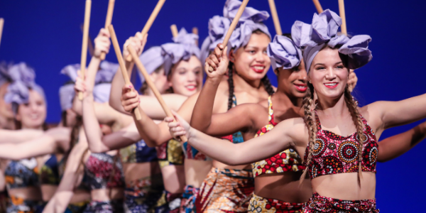 group of students dancing on stage in a line holding drumsticks and wearing headwraps